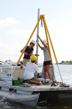 A yellow tripod on a boat supports soil sampling equipment. Two researchers are standing and one kneeling, all hands on the metal sampling tube.