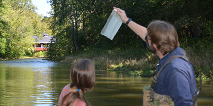 Two researchers wearing wading boots stand in a creek. The researcher on the left is holding up a water sample in a plastic container. In the background are woods and a covered bridge.