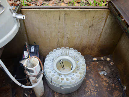 Inside an cement enclosure, we see an automated sampler on the left, a PVC pipe next to it, and a carousel of 24 water samples. 