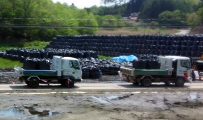 Wooded hills are behind tall stacks of black plastic bags, Two open-bed trucks transport more bags on a dirt road. In the foreground is tan-colored soil with a few puddles.