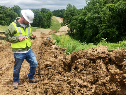 A reclamation site worker, wearing a hard hat and yellow safety vest, looks down at a chunk of soil he's holding on a spade. Behind him, the rolling soil path of the reclaimed pipeline area goes through trees on each side.