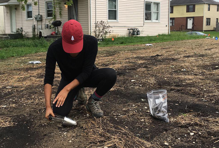 A person in a red hat is collecting a soil sample from bare ground near a house. There is a plastic bag holding some soil.